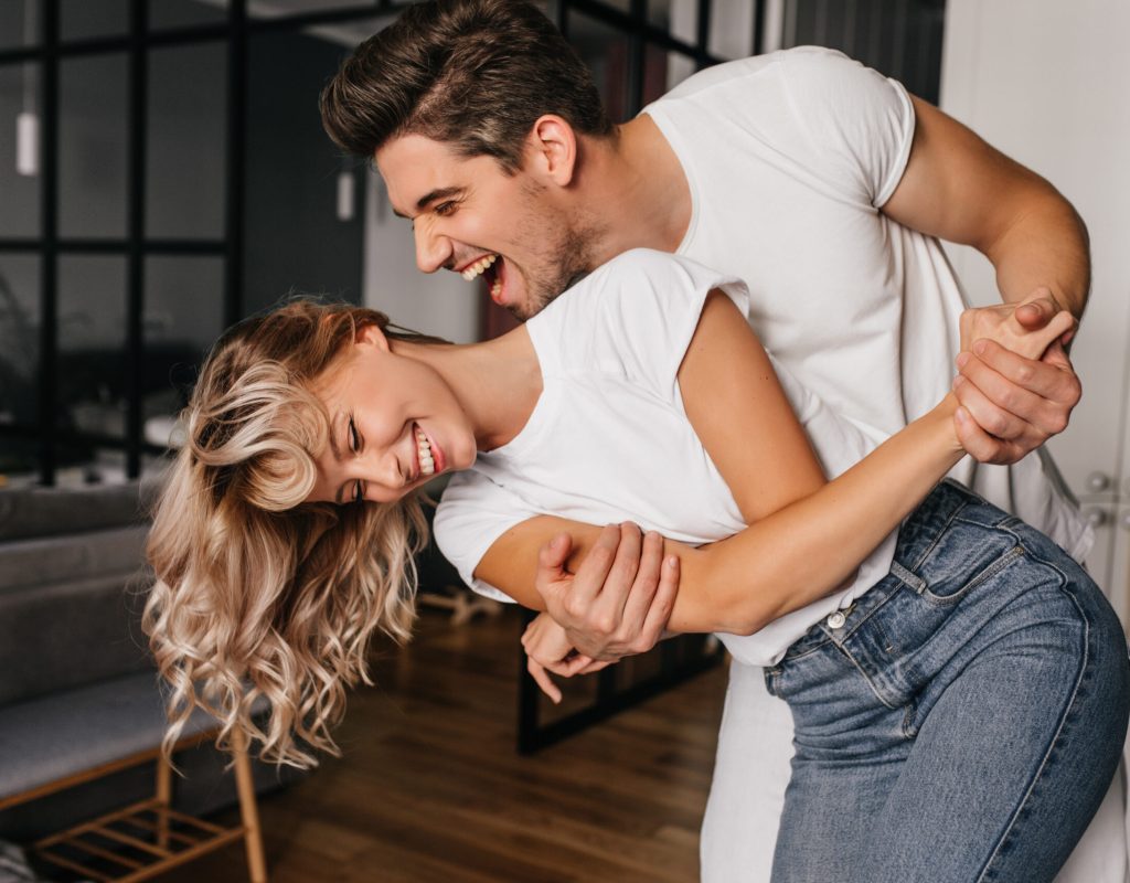 Wonderful girl in white t-shirt dancing with boyfriend. Indoor photo of winsome lady fooling around at home with husband.